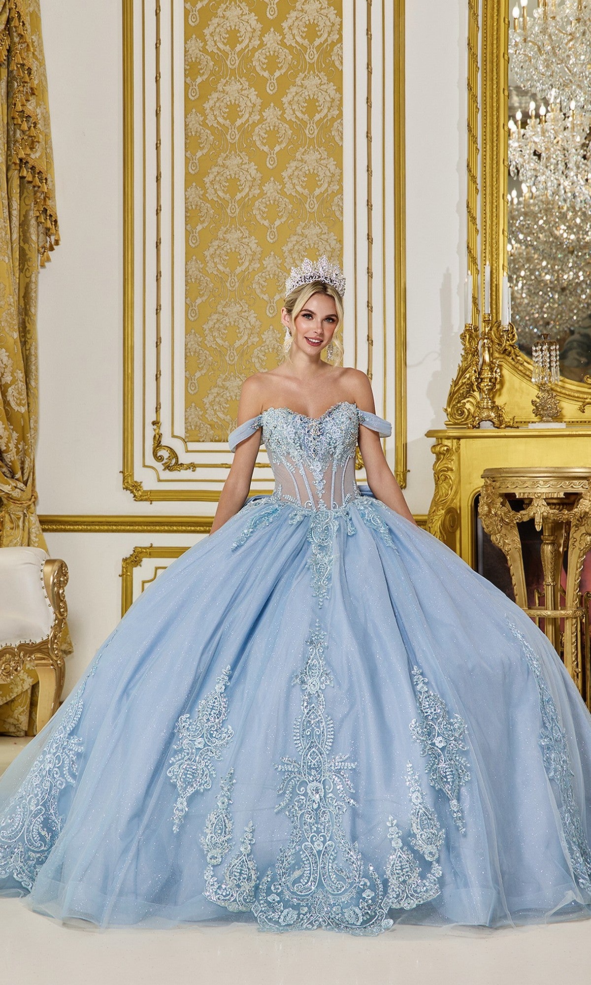 A woman wears the Quinceanera Dress 5063 by Dancing Queen in dusty blue, featuring a sheer corset bodice and glitter tulle, as she stands in an opulent room with gold accents, intricate walls, and a chandelier.